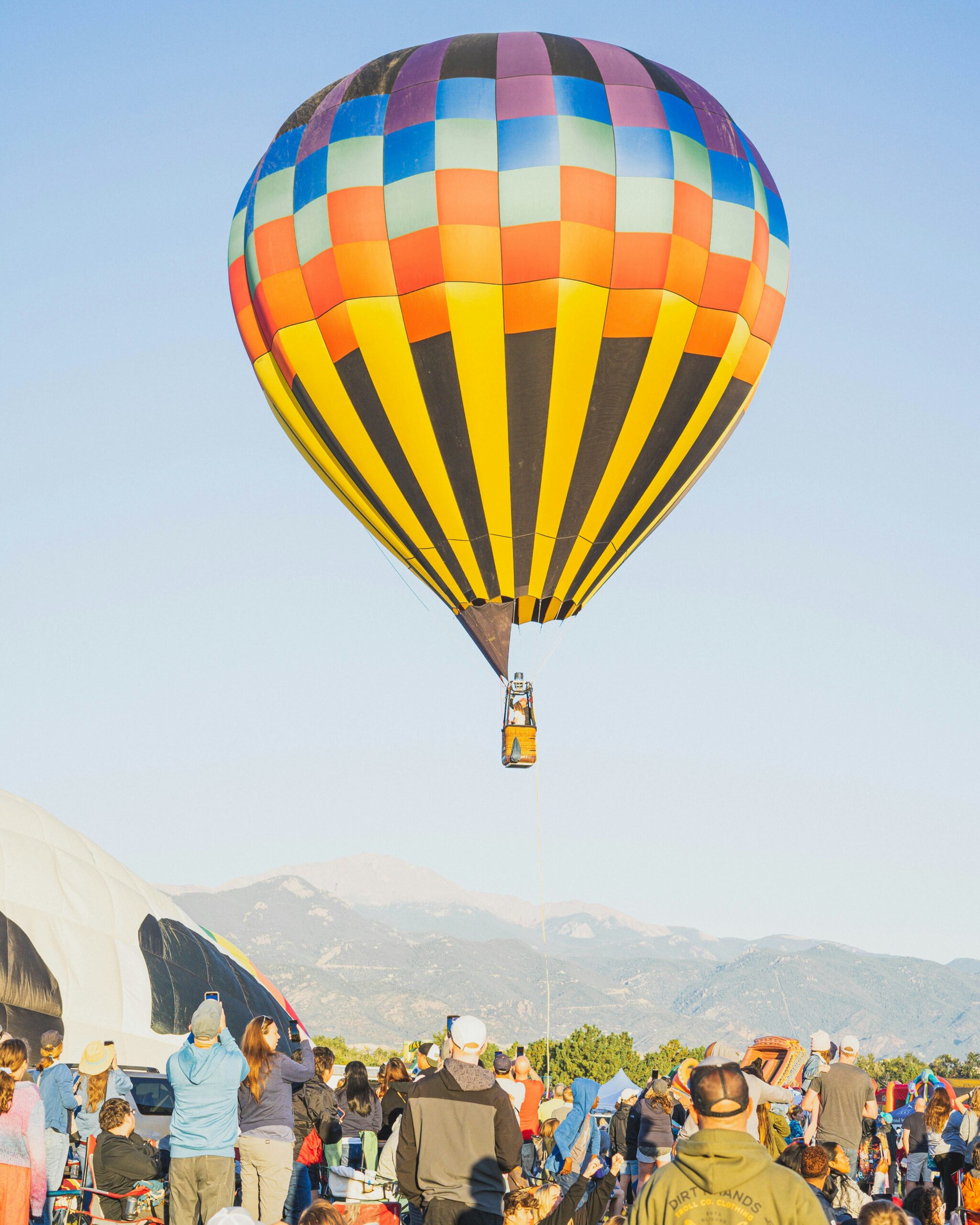 Eine bunt gemischte Gruppe von Menschen blickt einem startenden Heißluftballon hinterher beim Start in einen sonnigen Himmel. 